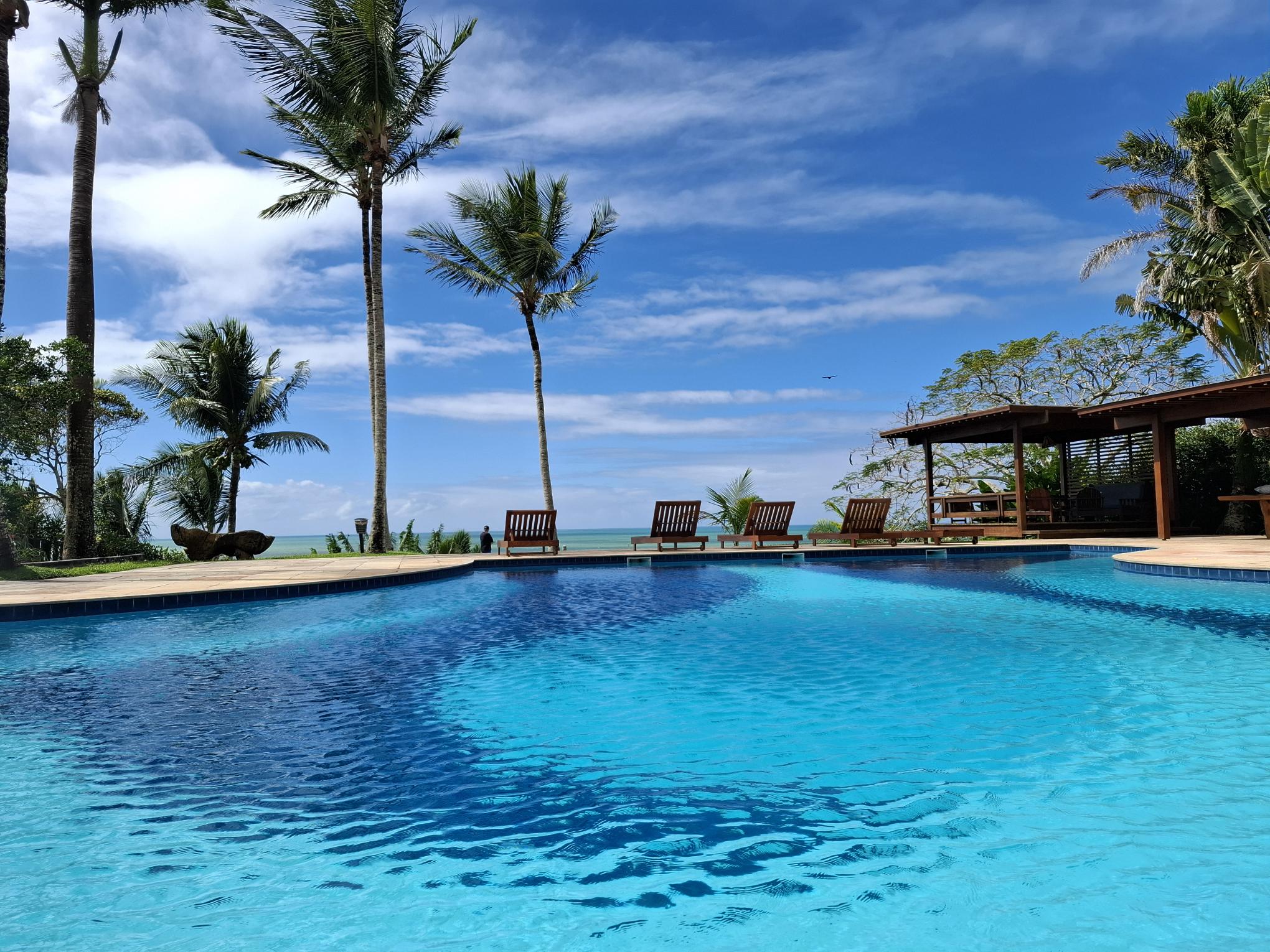 Piscina de uma pousada em Arraial d'Ajuda com vista para o mar, cercada por coqueiros sob um céu azul com nuvens, transmitindo tranquilidade e contemplação.